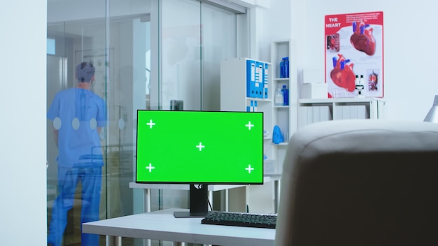 Компьютер с копией пространства в больнице и помощник идет в лифт. Рабочий стол с макетом пустого зеленого экрана изолировал пространство, доступное специалисту по медицине в кабинете клиники.