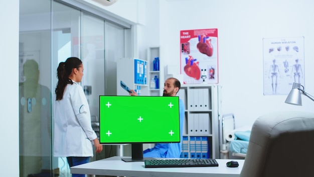 Компьютер с зеленым экраном в больничном шкафу и врач, проверяющий рентгеновский снимок пациента. Рабочий стол со сменным экраном в медицинской клинике, пока врач проверяет рентгенограмму пациента для постановки диагноза.