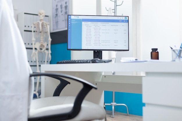 건강 검진 중에 빈 의사 사무실의 테이블에 서 있는 화면에 의료 환자 파일이 있는 컴퓨터. 전문적인 모던 가구를 갖춘 병원 사무실. 의료 지원