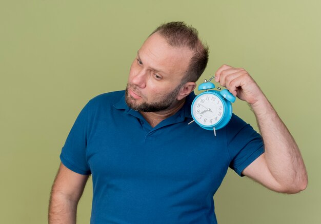 Concentrated adult slavic man holding alarm clock near ear tilting head to side looking down 