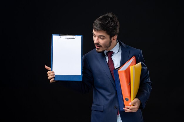 Concentrated adult in suit holding several documents and showing one of them on isolated dark wall