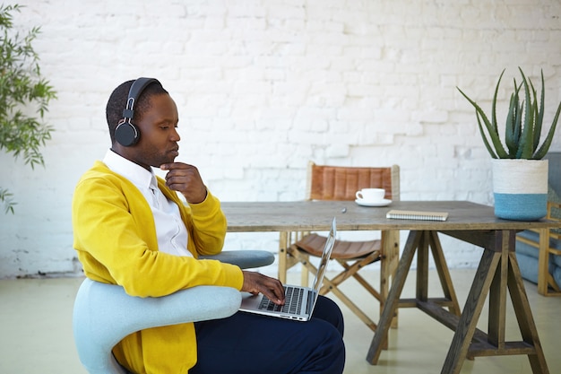 Concentrated African student wearing yellow cardigan and wireless headset studying online using wifi on generic laptop. Serious focused dark skinned freelancer working remotely on portable computer