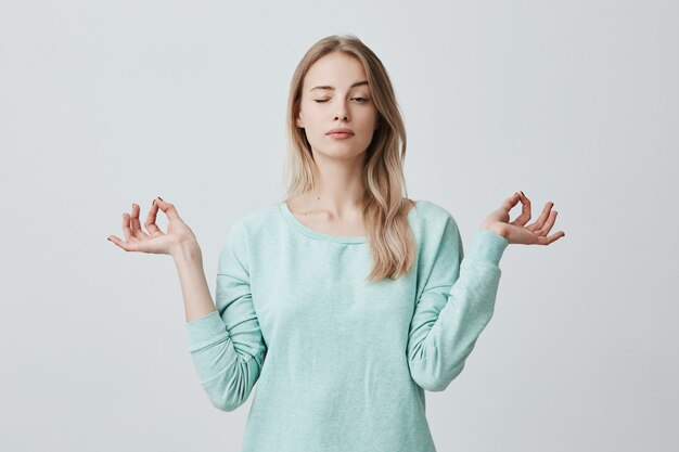 Concentrated attractive female with long dyed hair dressen in blue stands in lotus pose, meditates and enjoys peaceful atmosphere, closes eyes, tries to relax after hard working day. Mudra gesture