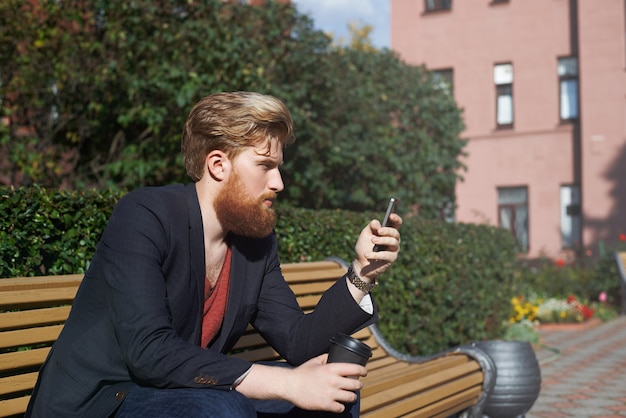 Concentrated bearded hipster using smarphone while sits on a bench