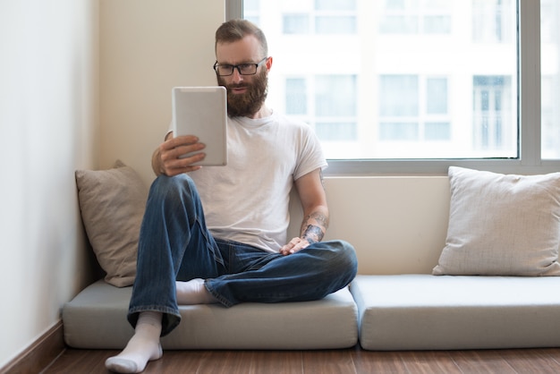 Concentrated bearded man sitting on pillow and using tablet