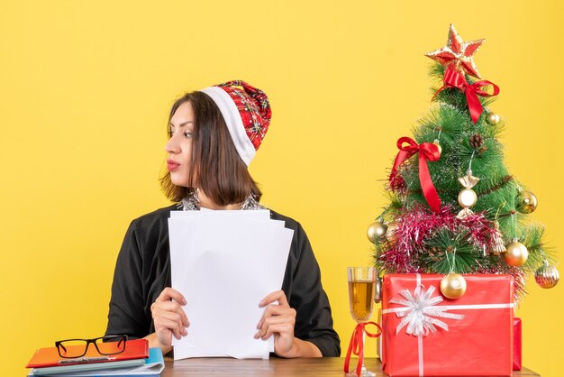 Concentrated business lady in suit with santa claus hat and new year decorations holding documents and sitting at a table with a xsmas tree on it in the office