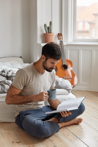 Concentrated businessman enjoys literature hobby, rests with book in bedroom, sits in lotus pose on floor, holds cup