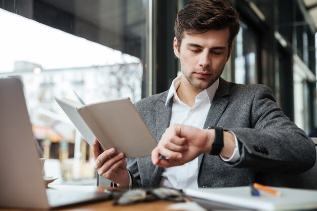 Concentrated businessman sitting by the table in cafe with laptop computer while holding book and looking at wristwatch