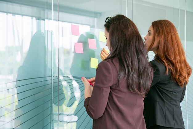 Concentrated businesswomen sticking notes on glass wall in conference room