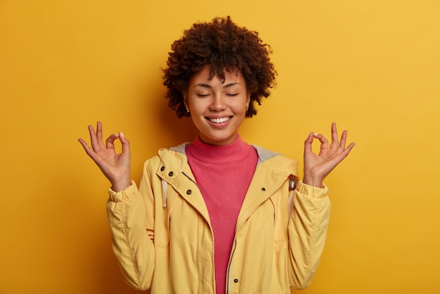 Concentrated cheerful curly woman keeps both hands in okay gesture, meditates indoor, has eyes closed, wears yellow anorak