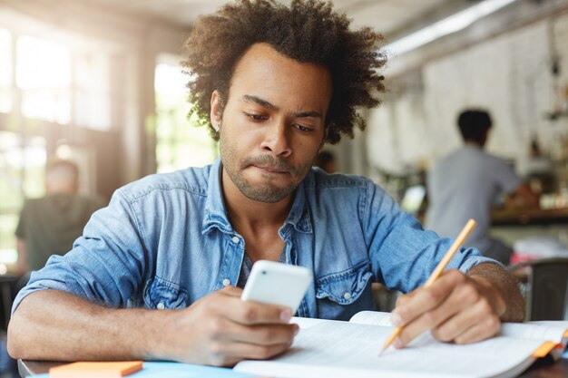 Concentrated dark-skinned college student with Afro hair doing homework, sitting at canteen table with textbook and copybook, making small break to read text message on his electronic device