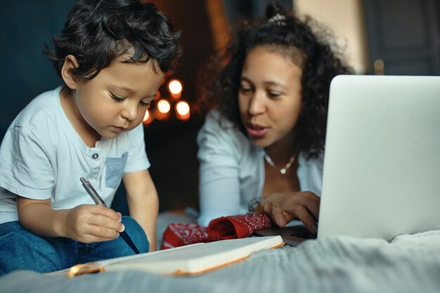 Concentrated dark skinned little boy learning alphabet, writing down letters in copybook, sitting on bed with his young mother using portable computer for remote work.