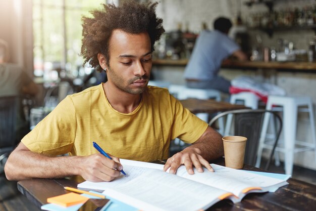 Concentrated dark-skinned male with African hairstyle and bristle wearing casual clothes writing notes in copybook and reading books while sitting at wooden table in cafeteria and drinking coffee.