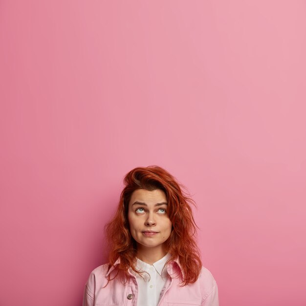 Concentrated European woman with ginger hair, healthy skin, looks upwards, wears pink clothes