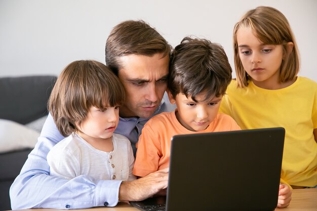画面を見ている集中父と真面目な子供たち。ノートパソコンの画面に入力する白人の中年のお父さんと彼の作品を見ている子供たち。父性、子供時代、デジタル技術の概念