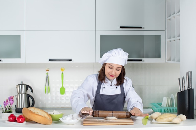 Free photo concentrated female commis chef in uniform standing behind table preparing pastry in the white kitchen