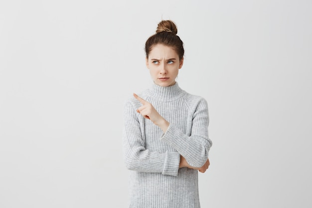 Concentrated female looking to side and showing with index finger. Woman with hair in bun having strict facial expressions frowning expressing don't like this.