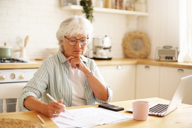 Concentrated female pensioner wearing eyeglasses focused on financial papers while paying bills online using laptop, holding pencil, making notes. People, technology, finances and domestic budget