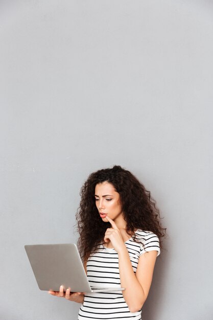 Concentrated female student with curly hair standing with notebook in hands studying hard or reading interesting ebook over grey wall