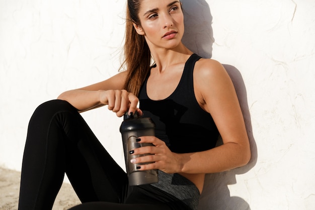 Free photo concentrated fitness lady sitting outdoors drinking water.