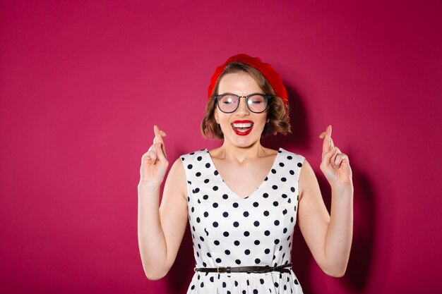 Concentrated ginger woman in dress and eyeglasses praying with crossed fingers and closed eyes over pink