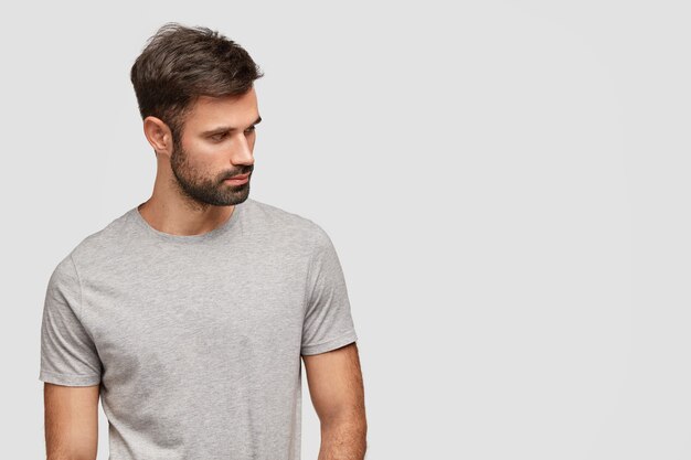 Concentrated handsome unshaven young man foused aside, has muscular body, dressed in casual grey t shirt