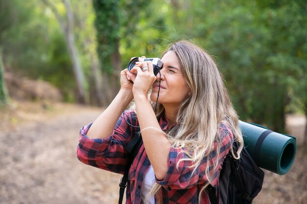 Concentrated lady shooting landscape and walking with backpack. Female tourist exploring nature, holding camera and taking photo. Tourism, adventure and summer vacation concept