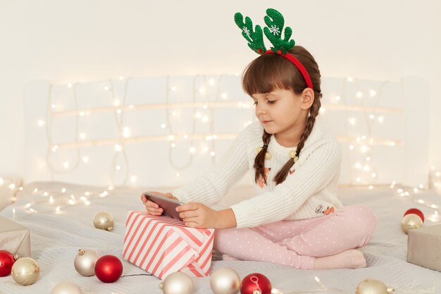 Сосредоточенная маленькая очаровательная девочка в белом повседневном свитере сидит на кровати с мобильным телефоном, смотрит мультфильмы или видео, позирует в праздничной спальне с шарами, подарочной коробкой и гирляндами.