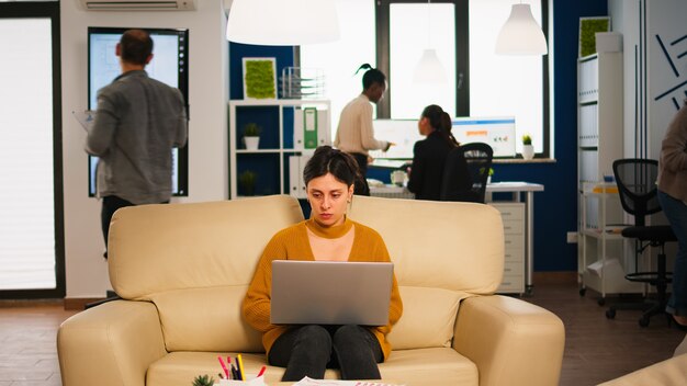 Concentrated manager reading mails typing on laptop sitting on couch in busy start up office while diverse team analyzes statistics data in background. Multiethnic team talking about project