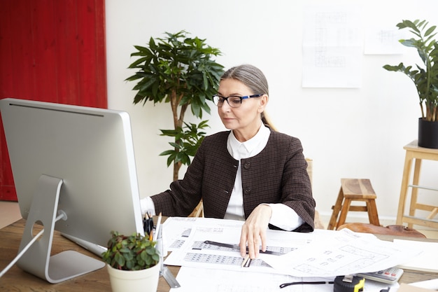 Concentrated mature female architect making drawings, comparing them with measures on computer. skilled woman engineer filing in electronic specification, looking at screen with focused expression