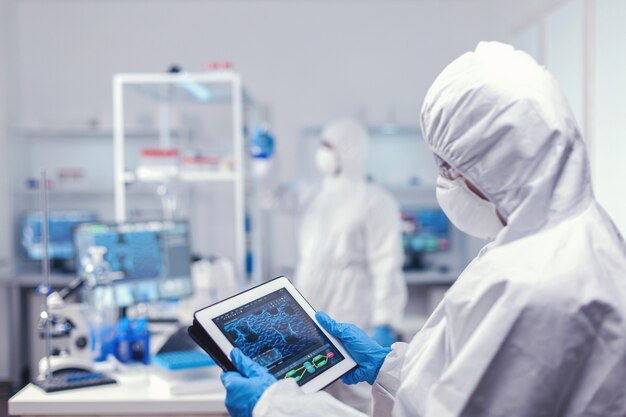 Concentrated medical researcher using digital tablet dressed in protective suit against infection with coronavirus. Team of scientists conducting vaccine development using high tech technology for res