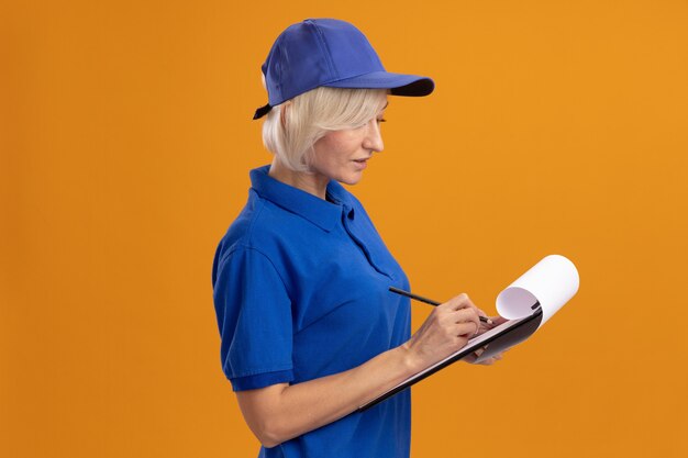 Concentrated middle-aged blonde delivery woman in blue uniform and cap standing in profile view writing with pencil on clipboard isolated on orange wall with copy space