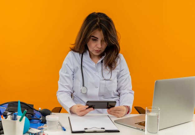의료 가운과 청진기를 착용하고 의료 도구 클립 보드와 노트북을 들고 고립 된 휴대 전화를보고 책상에 앉아 집중된 중년 여성 의사