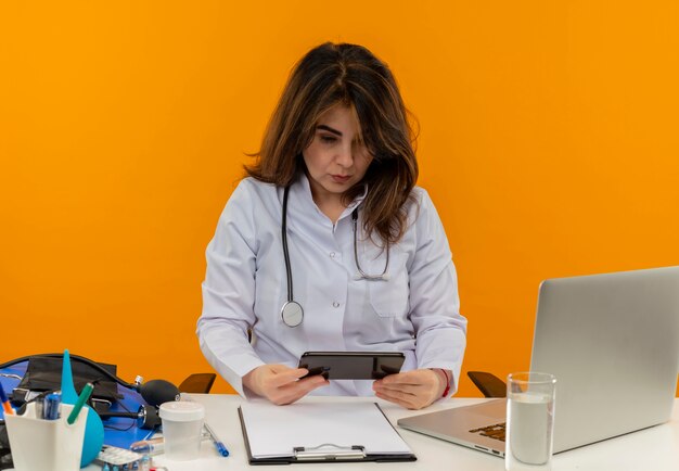 Concentrated middle-aged female doctor wearing medical robe and stethoscope sitting at desk with medical tools clipboard and laptop holding and looking at mobile phone isolated