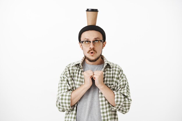 сосредоточенный игривый смешной европейский мужчина с бородой в очках и шапочке, держащий ладони возле груди, напряженно и обеспокоенно открывает рот и держит бумажный стаканчик с кофе на голове, балансирует