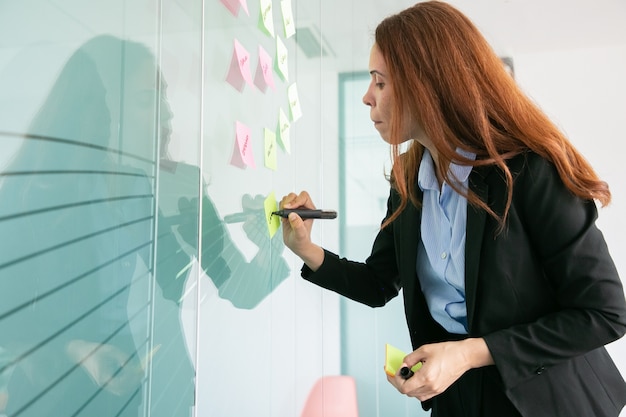 Foto gratuita donna di affari dai capelli rossi concentrata che scrive sull'adesivo con l'indicatore