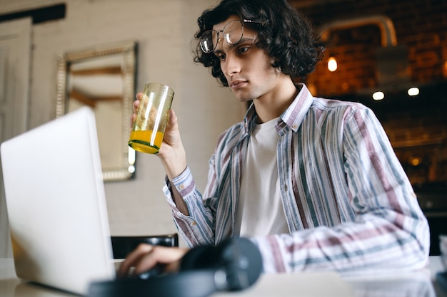 Concentrated serious young businessman in casual shirt working from home, sitting at desk in front of open laptop, checking email in the morning, drinking healthy fresh orange juice for breakfast