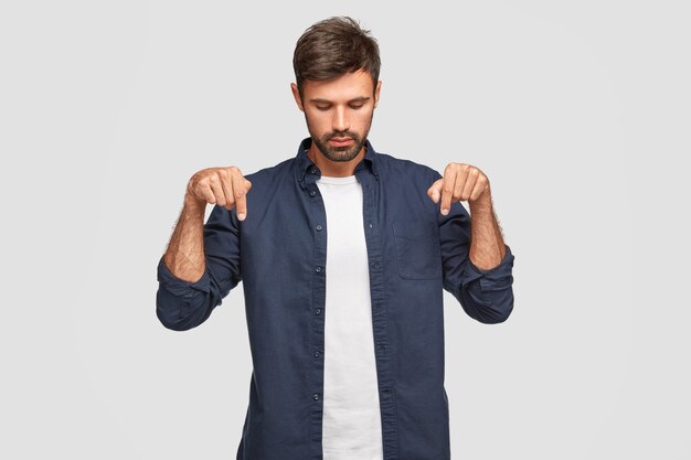 Concentrated serious young European male with appealing appearance focused down, indicates with both index fingers, has beard and mustache, wears elegant-shirt, isolated over white wall