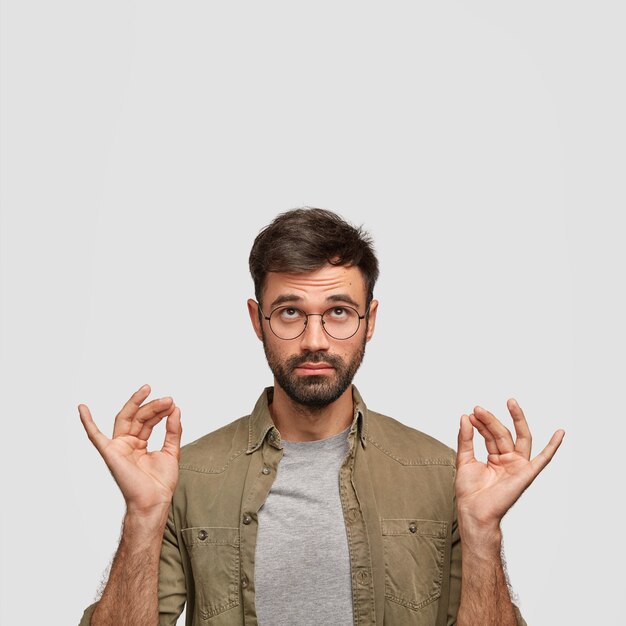 Concentrated unshaven guy believes in good fortune, makes mudra sign with both hands, meditates after work