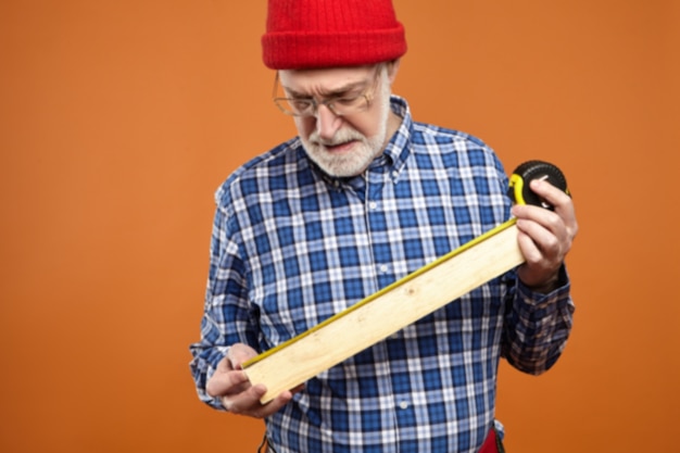 Concentrated unshaven retired carpenter wearing red knitted hat, tool belt and plaid shirt making furniture, holding wooden plank and measuring tape. Craft, job, occupation, age and retirement