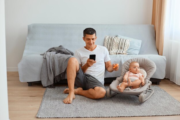 白いtシャツを着て、ロッキングチェアに幼児の娘と一緒に床に座って、携帯電話を使用して、彼の幼児の子供と遊んでいる集中した若い大人の白人男性。