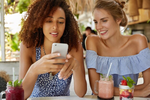 Concentrated young African American woman reads news online on cell phone and her female friend being curious, looks at screen, has positive expression. Relaxed multiethnic women rest in restaurant