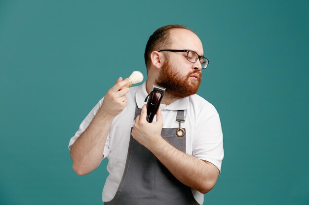 Concentrated young barber wearing uniform and glasses turning head to side with closed eyes holding hair trimmer and shaving brush trimming his own face isolated on blue background