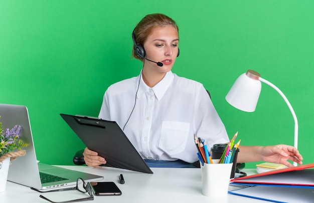 Foto gratuita concentrato giovane bionda call center ragazza che indossa l'auricolare seduto alla scrivania con strumenti di lavoro tenendo appunti toccando e guardando la cartella