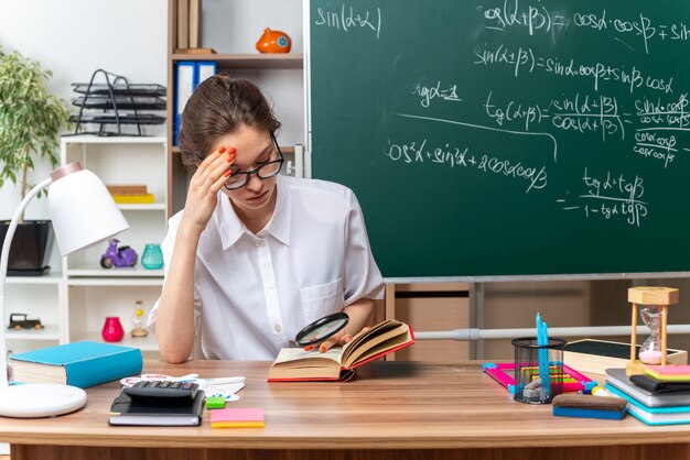 Concentrated young blonde female math teacher wearing glasses sitting at desk with school tools looking at book through magnifying glass keeping hand on forehead in classroom