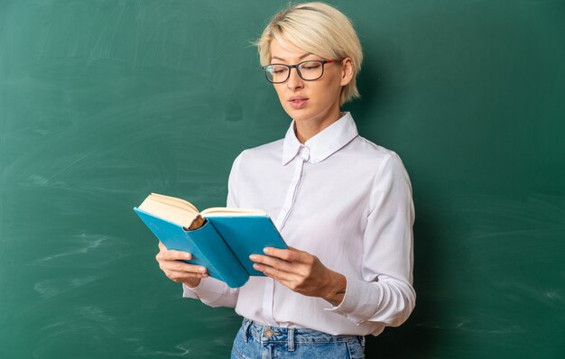 책을 들고 칠판 앞에 서 있는 교실에서 안경을 쓴 집중된 젊은 금발 여교사