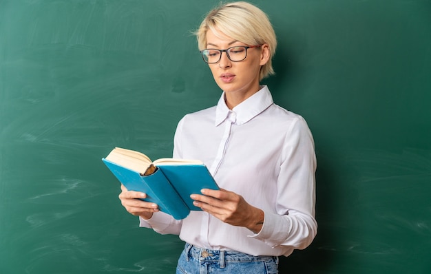 concentrated young blonde female teacher wearing glasses in classroom standing in front of chalkboard holding and reading book