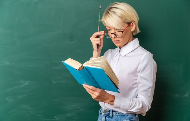 Concentrated young blonde female teacher wearing glasses in classroom standing in profile view in front of chalkboard holding pointer stick and reading book grabbing glasses with copy space