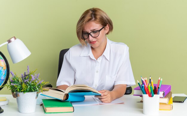オリーブグリーンの壁に分離された本を読んで学校のツールと机に座って眼鏡をかけて集中若いブロンドの学生の女の子
