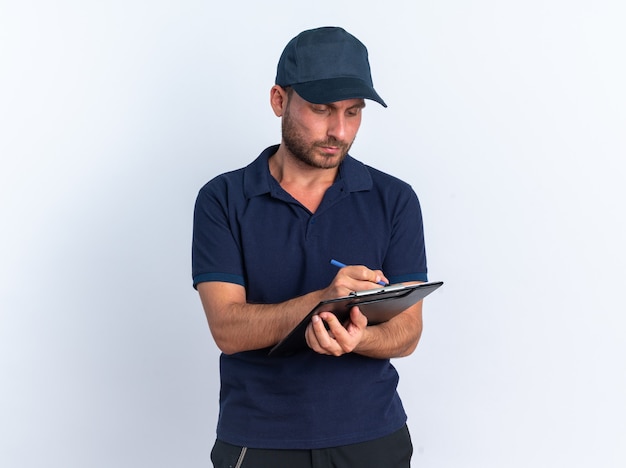 Concentrated young caucasian delivery man in blue uniform and cap writing with pen on clipboard isolated on white wall with copy space